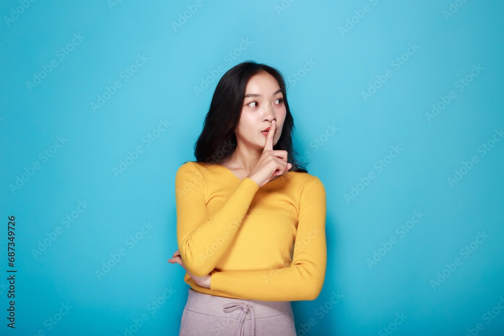 An Asian woman poses to signal silence while standing in front of a light blue background. An Asian woman poses to signal silence while standing in front of a light blue background.
