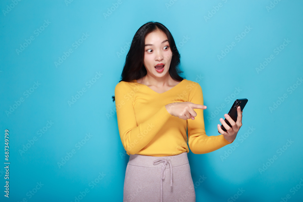 Young happy woman showing point fingers isolated while holding a cell phone in the other hand, Standing and pointing happily, light blue back ground. Young happy woman showing point fingers isolated while holding a cell phone in the other hand, Standing and pointing happily, light blue back ground.
