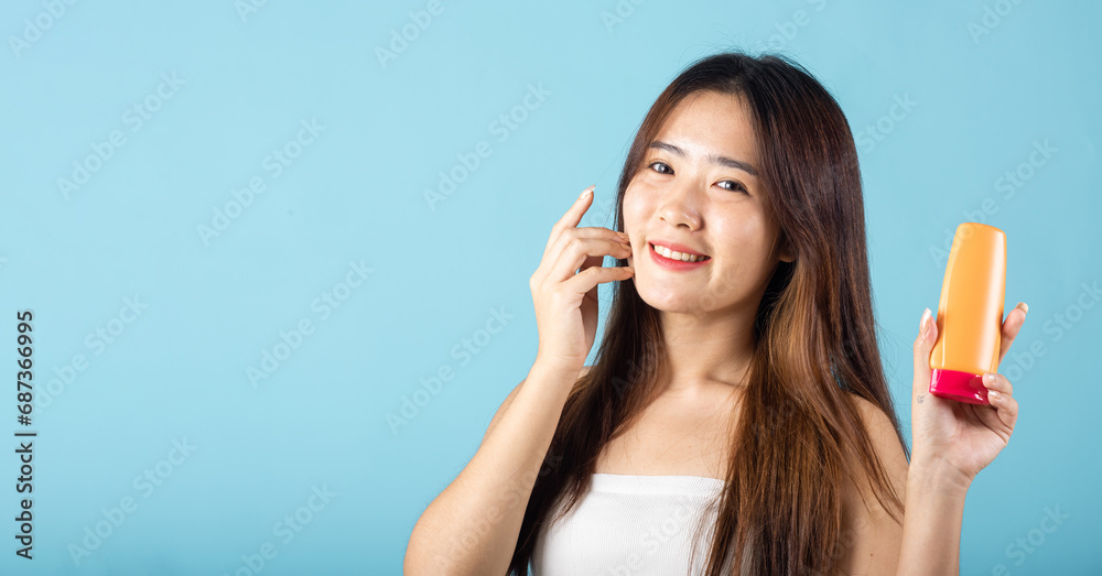 Asian young woman holding sunscreen lotion cream with hand touching on face studio shot isolated on blue background, Happy smiling teen female present sun protection skin care