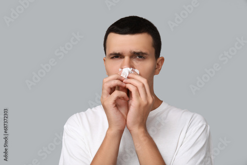 Ill young man with tissue blowing nose on grey background