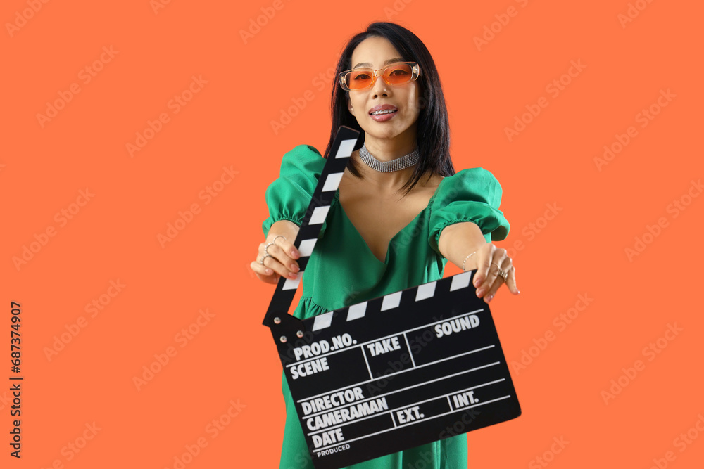 Beautiful stylish Asian woman in sunglasses with clapperboard on orange background