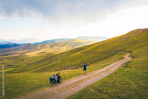 Aerial inspirational view excited joyful caucasian male cyclist standing by red touring bicycle looking to scenic mountains background and stand confident carefree with hands up