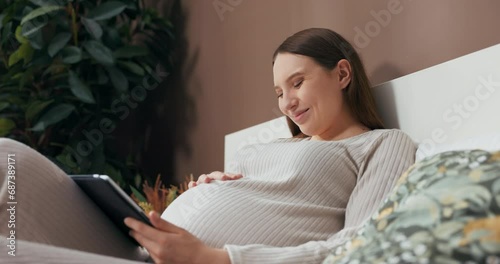 Dreamy alluring pregnant woman dressed in a stylish and comfortable outfit, sitting on a bed, holding a tablet, and admiring photos with interest. photo