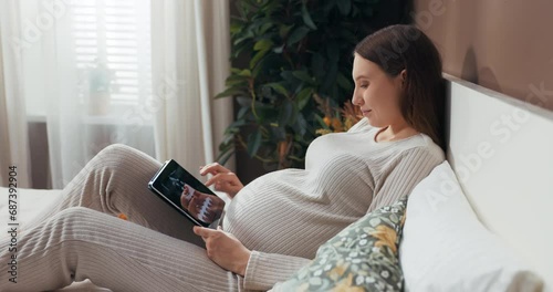 Attractive pregnant woman in a stylish and comfortable outfit sits on a bed holding a tablet, scrolling through photos with a curious expression. photo