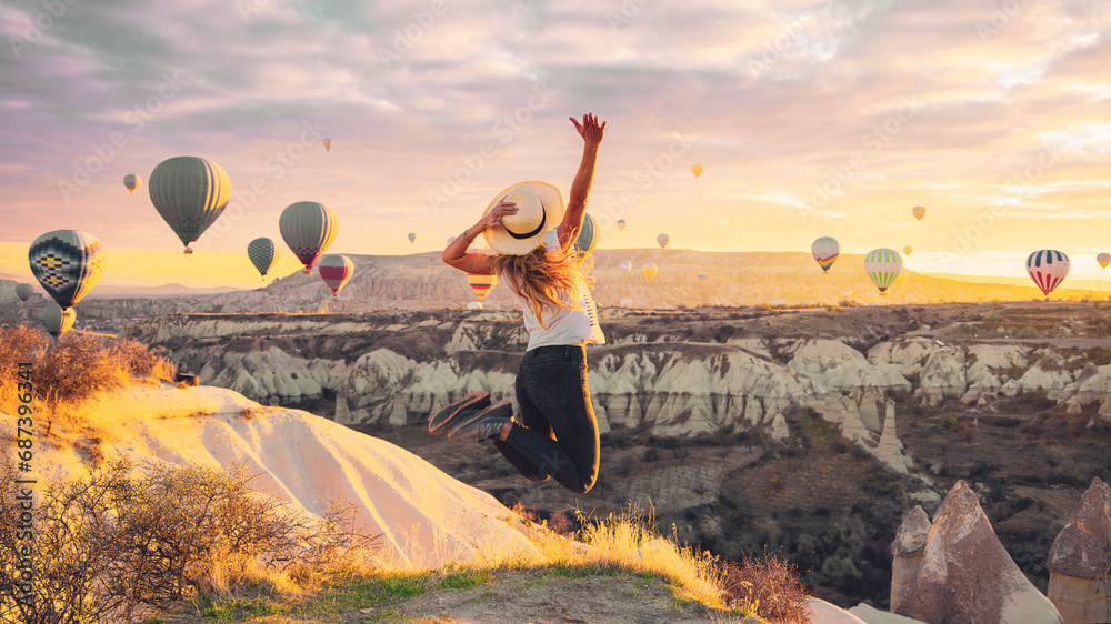 Young female tourist jumping in the air with hot air balloons background at sunrise in Cappadocia, Turkey - obrazy, fototapety, plakaty 
