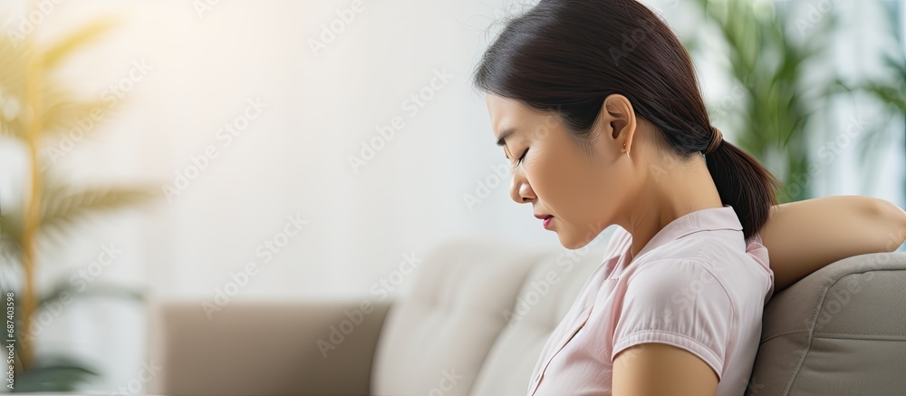 Asian woman with posture issue experiences back pain while sitting on the sofa, massages shoulders for relief and to relax muscles. Asian woman with posture issue experiences back pain while sitting on the sofa, massages shoulders for relief and to relax muscles.