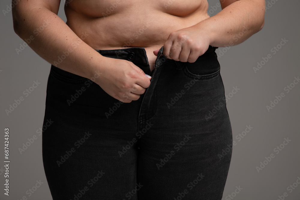 Overweight woman trying trying to button up her pants. Plus size model in tight black jeans on grey background.
