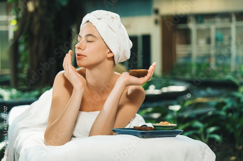 Beautiful caucasian women in white towel smells herbal homemade facial mask while lie on spa bed surrounded by outdoor natural environment. Relaxing and chilling concept. Tranquility.