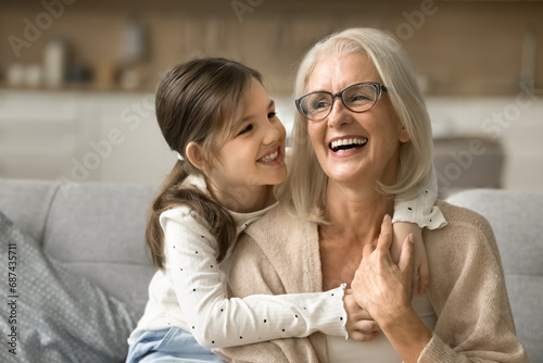 Happy blonde grandma and cheerful granddaughter child playing active games on home sofa. Grandmother and grandkid enjoying funny leisure on family meeting, having fun, hugging on couch photo
