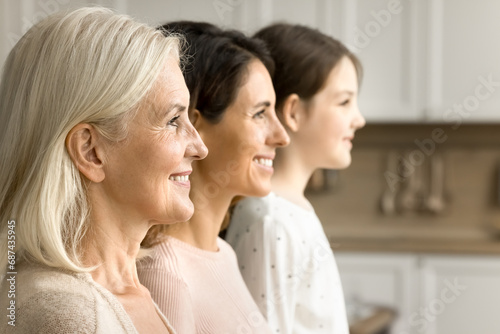 Cheerful blonde elderly grandmother looking forward away side shot with brunette daughter woman and grandkid girl standing in line in blurred background. Three female generations family portrait photo