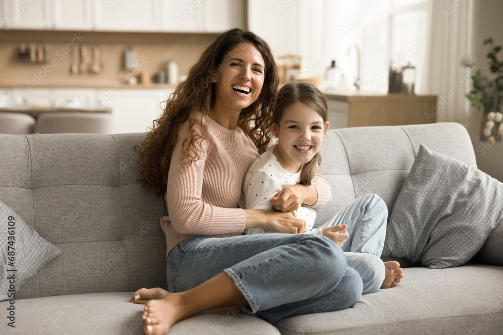 Cheerful pretty mom cuddling sweet daughter girl on couch, looking at camera with toothy smile, laughing, playing active games with kid, enjoying motherhood, childcare, playtime Cheerful pretty mom cuddling sweet daughter girl on couch, looking at camera with toothy smile, laughing, playing active games with kid, enjoying motherhood, childcare, playtime