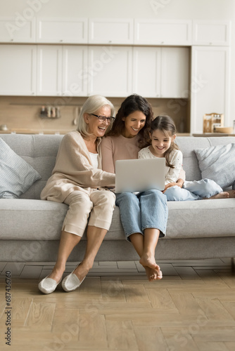 Positive excited grandmother, mom and little tween child using laptop on home couch together, watching online content, movie, enjoying Internet service, communication, digital entertainment