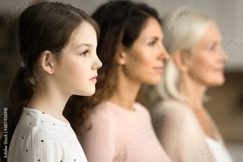 Side shot of serious little tween child girl standing in row with mom and grandmother in blurred background, looking forward away, posing for family portrait of three female generations photo