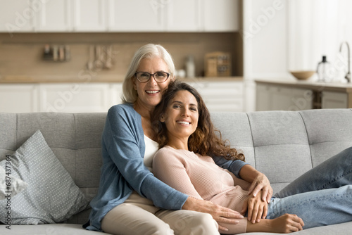 Happy blonde attractive mature mother hugging daughter woman on home couch, looking at camera, smiling, enjoying motherhood, close family relationship. Elderly parent and adult kid portrait photo