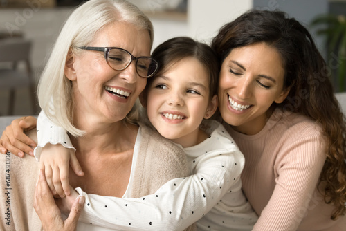 Happy girls and women of three family generations hugging with love, tenderness at home, feeling close family relations, bonding, smiling, laughing. Grandma, mom and kid girl meeting on mothers day photo