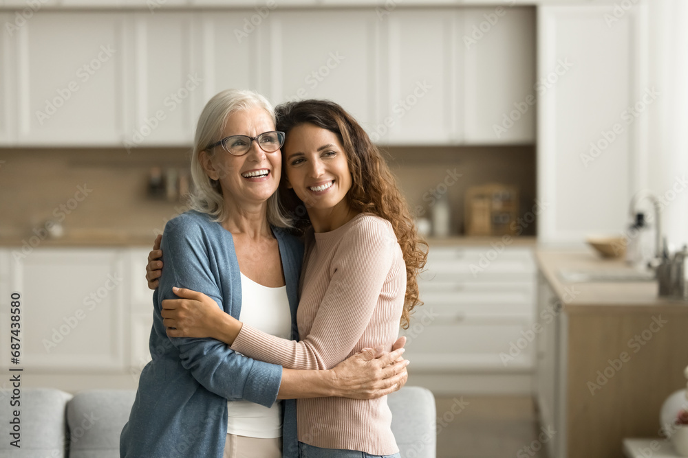 Cheerful dreamy elder mom and positive adult daughter woman hugging at home with love, support, looking away, smiling, thinking on family relationships, bonding, discussing mothers retirement plans Cheerful dreamy elder mom and positive adult daughter woman hugging at home with love, support, looking away, smiling, thinking on family relationships, bonding, discussing mothers retirement plans
