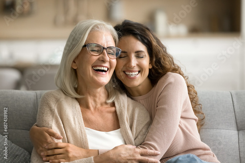 Happy excited senior mom and adult daughter woman having fun at home, hugging with heads touch, sitting on sofa, laughing with closed eyes, enjoying funny talk, leisure, family affection