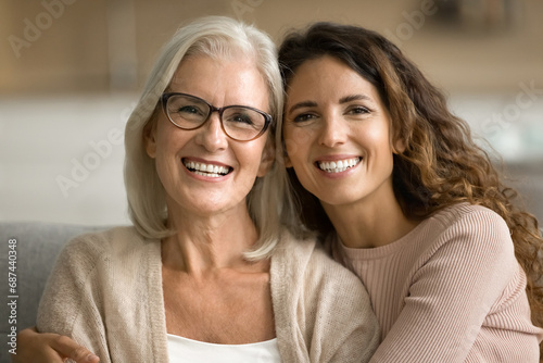 Happy blonde elderly mom and young daughter woman posing at home, looking at camera with toothy smiles, laughing, hugging, enjoying warm family relationship, bonding. Head shot portrait photo