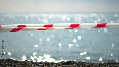 Red white warning tape barrier ribbon swinging in the wind across exotic sea beach background without people. No entry Red White caution tape. No holiday concept, delayed travel, no summer plans photo