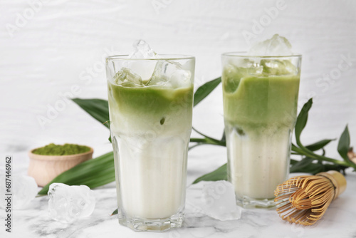 Glasses of tasty iced matcha latte, bamboo whisk and leaves on white marble table