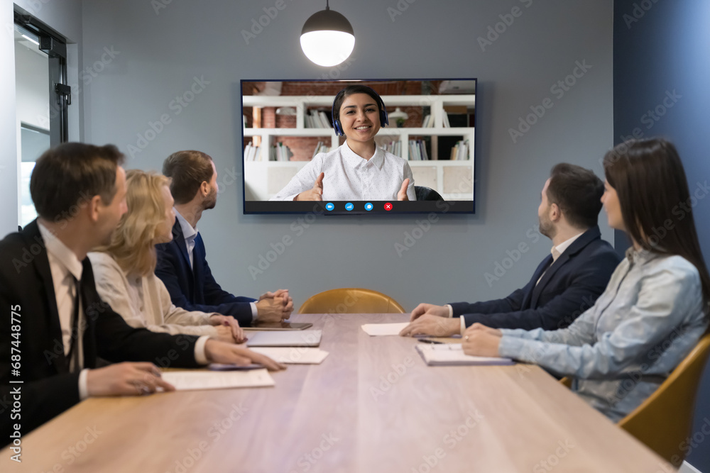 Indian woman leads on-line conference with diverse teammates sit at desk look at tv screen sit at desk in modern boardroom. HR manager interviewing Indian woman applicant answer questions at interview - obrazy, fototapety, plakaty 