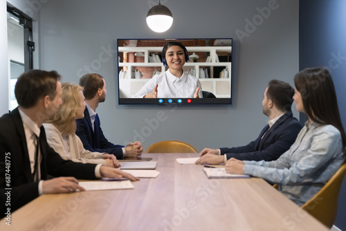 Indian woman leads on-line conference with diverse teammates sit at desk look at tv screen sit at desk in modern boardroom. HR manager interviewing Indian woman applicant answer questions at interview