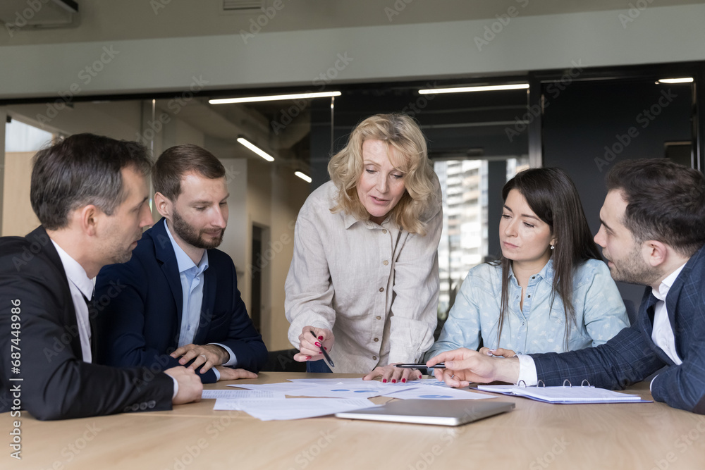 Focused group gathered together sit at desk in office boardroom at negotiations or morning briefing listening to mature female boss analysing sales statistics do paperwork, engaged in common project Focused group gathered together sit at desk in office boardroom at negotiations or morning briefing listening to mature female boss analysing sales statistics do paperwork, engaged in common project