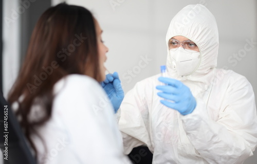 Portrait of doctor in protective white suit and mask take analysis for covid19 from patient. Infected woman sit with open mouth side view.