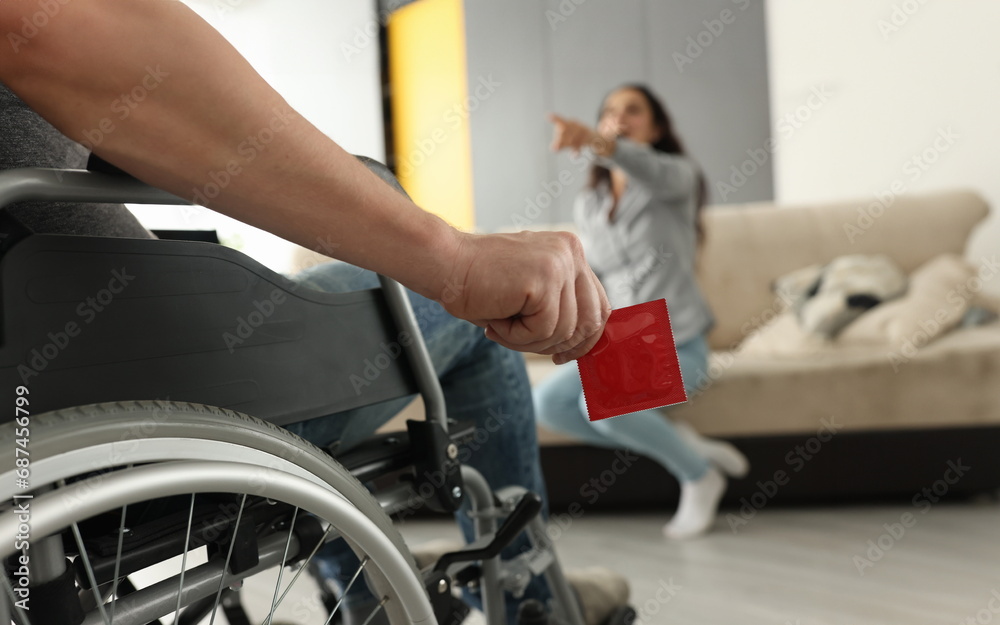 Obraz premium Woman sit on couch, point finger and laugh. Man in wheelchair hold red condom in his hand close up.