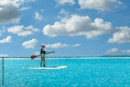 Cute little Caucasian child girl in hat sunglasses enjoying having fun standing on sup board surfing at Maldives tropical sea hot sunny day. Summer freedom leisure healthy sport recreation activities