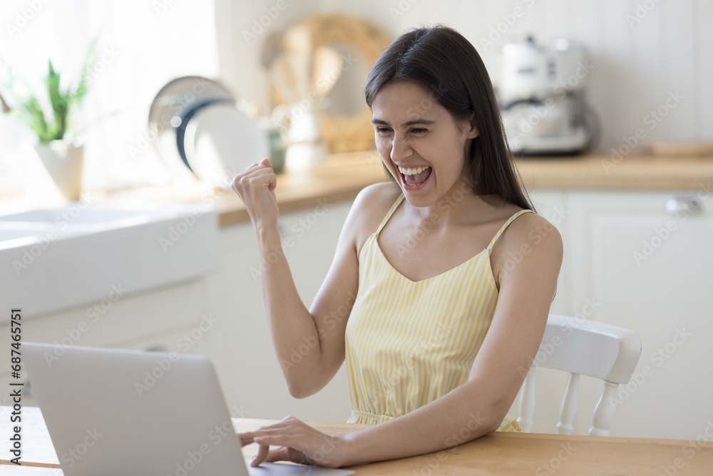 Excited woman sit at kitchen table use online application on laptop, looking at computer screen, read good news, celebrate win, rejoice success, show yes gesture, overjoyed lady enjoy luck, feel happy
