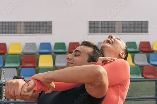Young heterosexual couple hugging each other on the sports field. Woman smiling energetically and grabbing the back of her partner's neck playing while both are smiling happily.