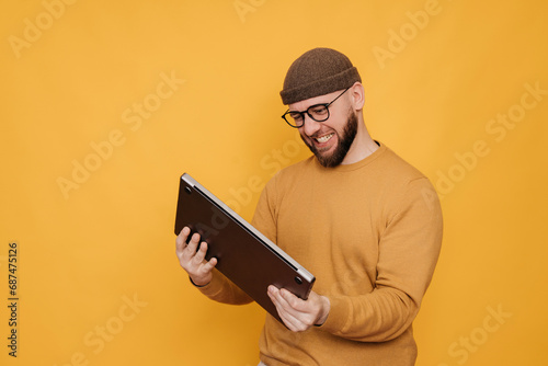 Angry young bearded  man in winter hat and casual sweater upset by laptop wants to break computer standing against  yellow studio background. Unhappy male annoyed by computer. photo