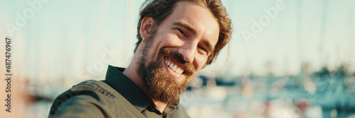 Closeup portrait of a smiling man with a beard chatting on the embankment, on a yacht background. Frontal close-up of happy young male hipster using mobile phone