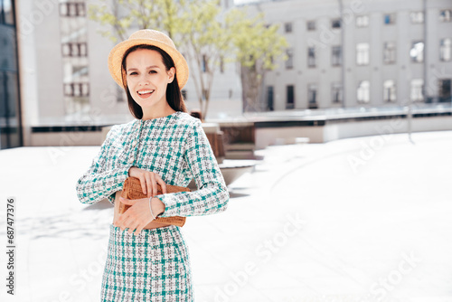 Young beautiful smiling woman in trendy summer green jacket and skirt clothes. Sexy carefree model posing in the street at sunset. Positive brunette female. Cheerful and happy. In hat, holds clutch