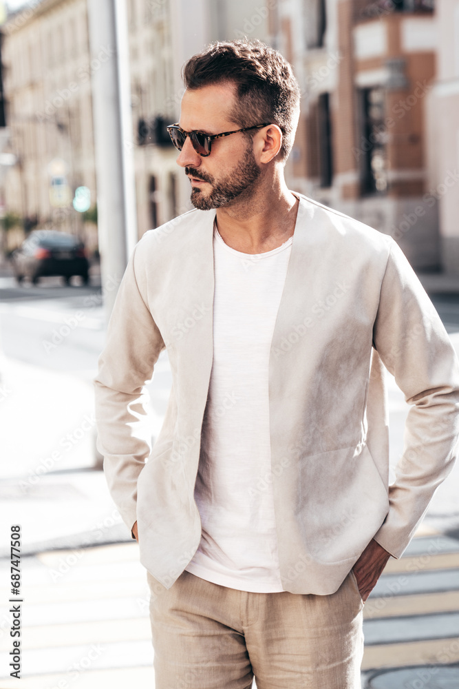 Obraz premium Handsome confident stylish hipster lambersexual model. Sexy modern man dressed in elegant white suit jacket. Fashion male posing in the street background in Europe city at sunset. In sunglasses