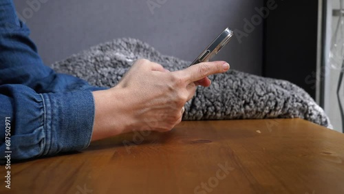 Female person typing message on the smartphone with thumb fingers. Side view medium shot of woman hands on wooden table on gray wall background, wearing navy blue jeans shirt, messaging on her phone. photo