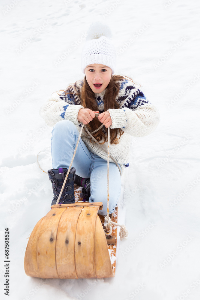 Fototapeta premium Selective focus vertical view of little blond girl in winter clothes making a funny face while sliding down a snowy slope in a vintage wooden sled