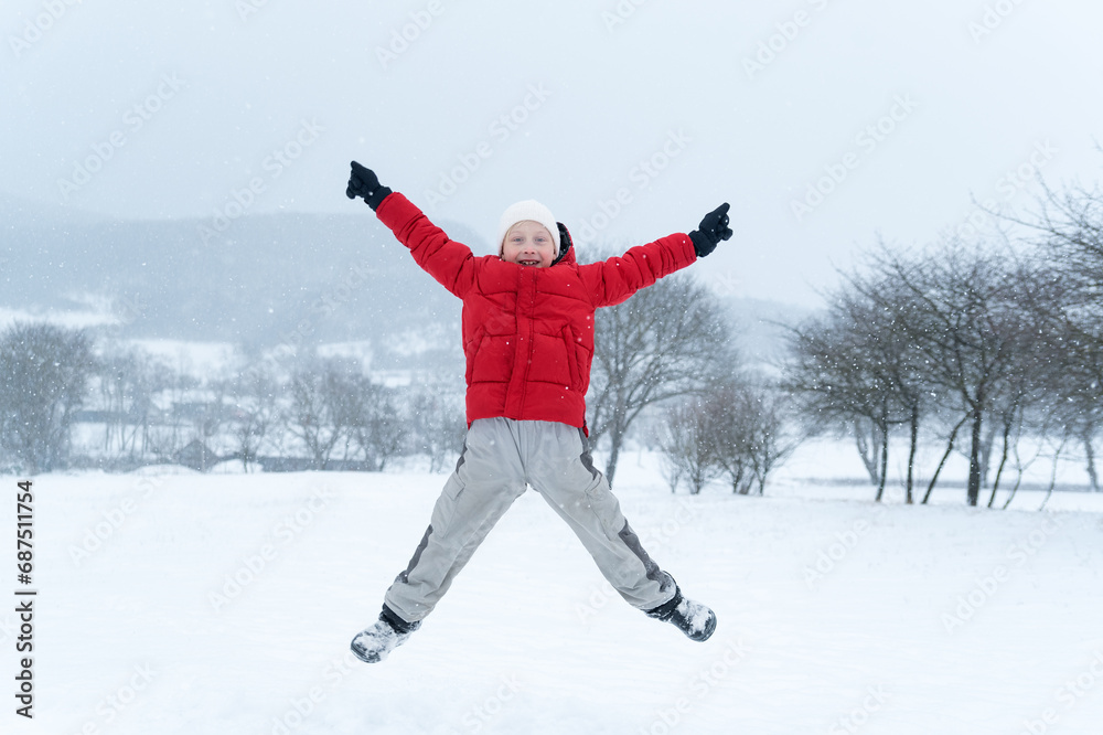 Child with his hands up and enjoys the winter holidays. Teenage boy in red jacket jumps during snowfall Child with his hands up and enjoys the winter holidays. Teenage boy in red jacket jumps during snowfall