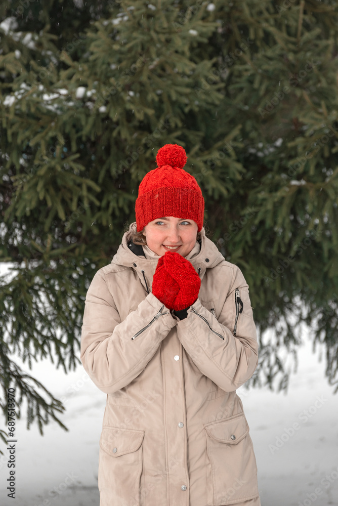 Smiling young woman in wool red hat and mittens warms up in the winter outdoors. Girl in comfortable clothes. Vertical frame
