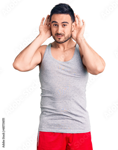 Young handsome man wearing swimwear and sleeveless t-shirt trying to hear both hands on ear gesture, curious for gossip. hearing problem, deaf