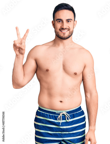 Young handsome man wearing swimwear smiling looking to the camera showing fingers doing victory sign. number two.
