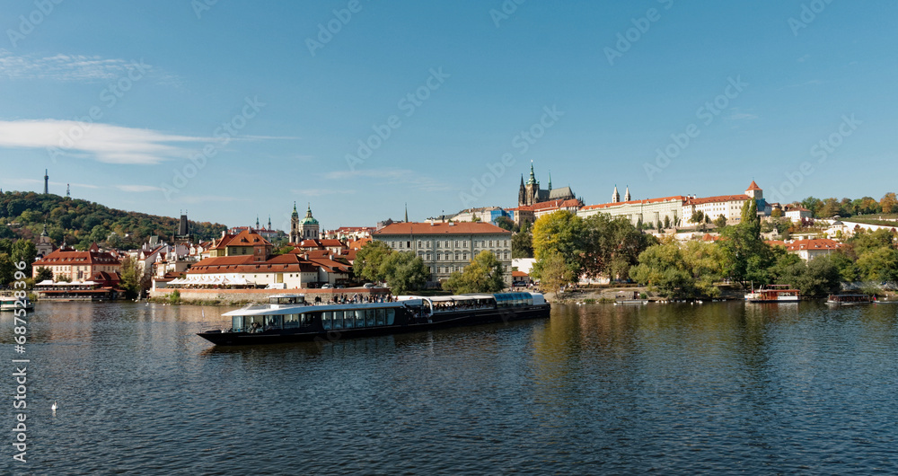 A boat is sailing on a river with a cityscape in the background. Pleasure boat on the Vltava pier in the fall. View of Prague.
