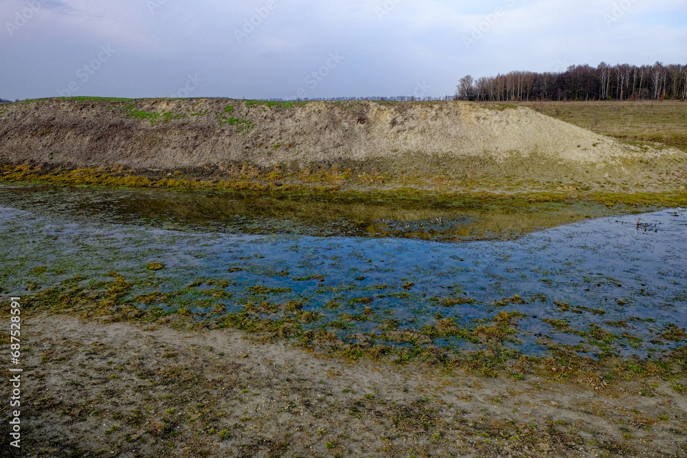 A landscape photo captures a shallow body of water with green algae covering the surface, surrounded by a dirt embankment. A landscape photo captures a shallow body of water with green algae covering the surface, surrounded by a dirt embankment.