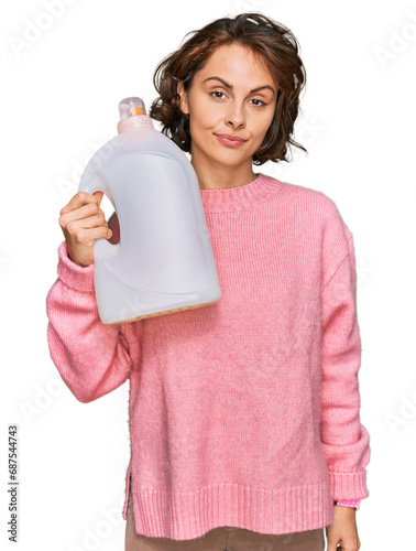 Young hispanic woman doing laundry holding detergent bottle thinking attitude and sober expression looking self confident