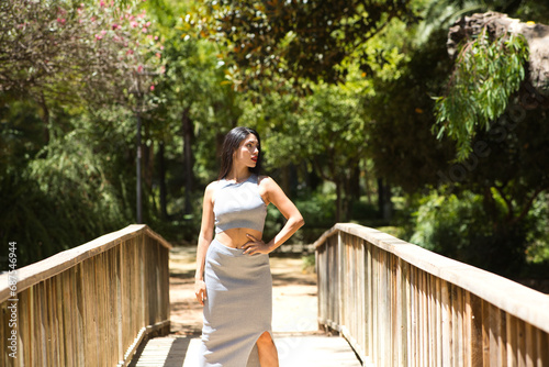 Latin woman, young and beautiful brunette walks on a wooden bridge in a park in seville. The woman is wearing an elegant grey dress and high heels and poses for the photo. Beauty and fashion concept.