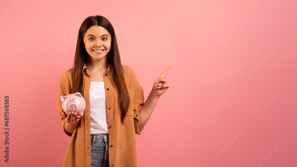 Smiling teen girl holding piggy bank and pointing at copy space Smiling teen girl holding piggy bank and pointing at copy space