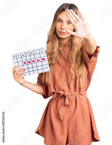 Beautiful caucasian woman with blonde hair holding heart calendar with open hand doing stop sign with serious and confident expression, defense gesture photo