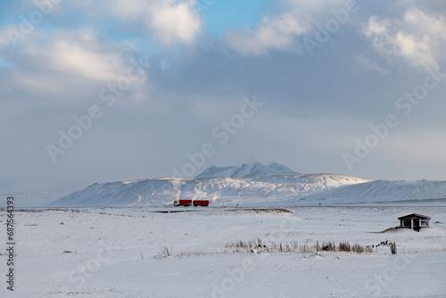 Panoramic view over snow covered landscape with snow covered mountains in skimming winter sunlight in background on Iceland with bright red truck as contrast on road in distance