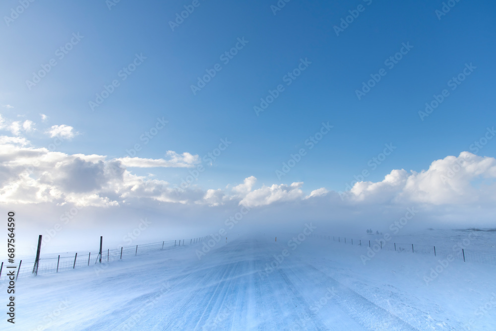 View over snow covered through agricultural snowy landscape with high winds blowing snow over road in Iceland with snow clouds in distance giving way to clear blue sunny sky View over snow covered through agricultural snowy landscape with high winds blowing snow over road in Iceland with snow clouds in distance giving way to clear blue sunny sky